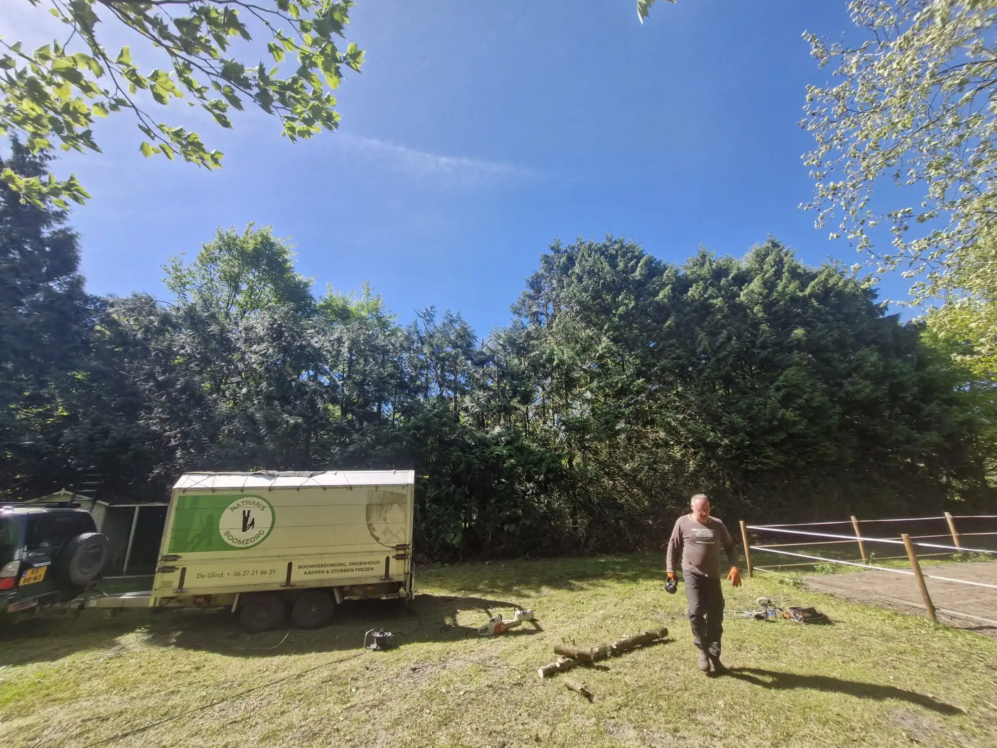 Nathan loopt over een gemaaid grasveld naar de camera, achter hem zijn aanhanger met logo en een rij bomen